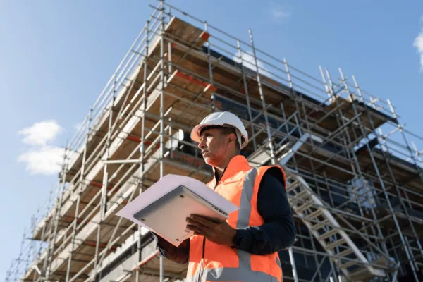 Construction worker in safety gear holding documents in front of scaffolding.