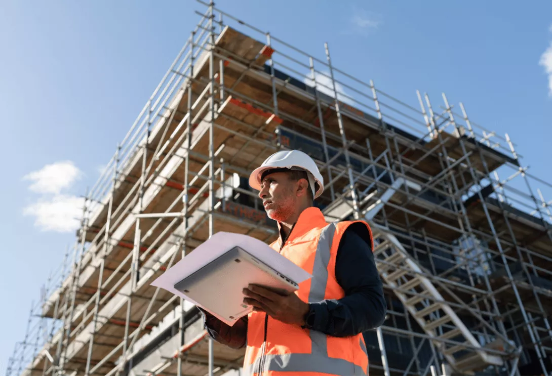 Construction worker in safety gear holding documents in front of scaffolding.
